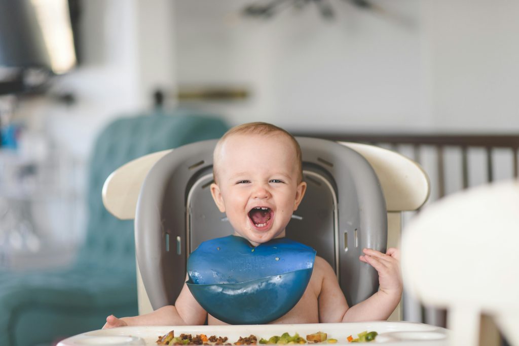 pexels-photo-9305088-9305088 Happy baby laughing in a high chair during mealtime, wearing a blue bib.